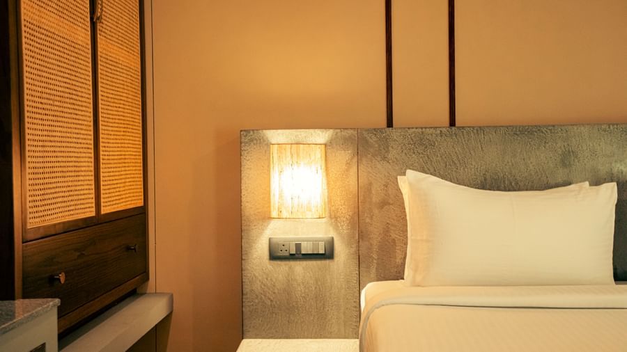 Close-up of a neatly made bed with a cushioned headboard, bedside light, and panelled wall at Fireflies Resorts, Kabini.