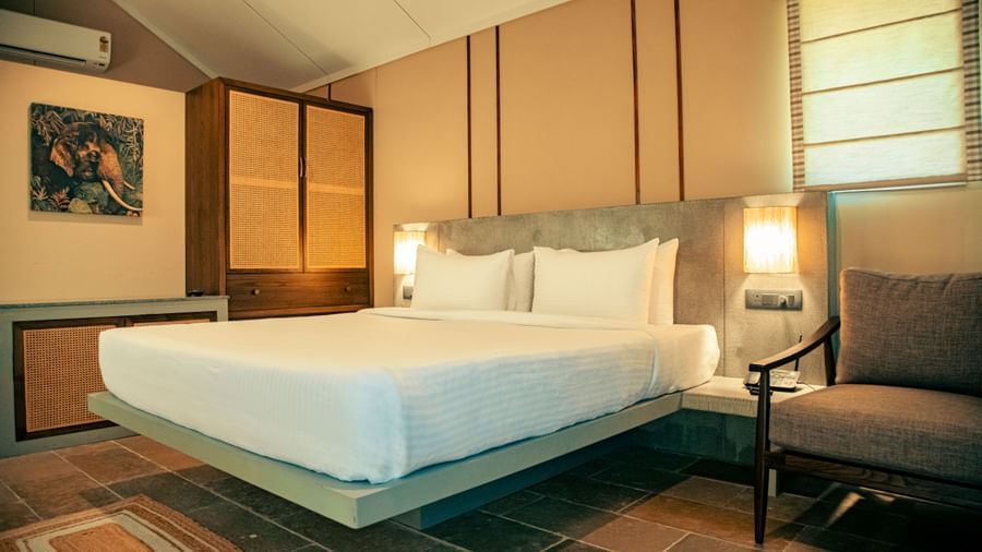 Room with wooden-panelled walls, king size bed with white bedding, two lamps, chair, and wall-mounted painting at Fireflies Resorts, Kabini.