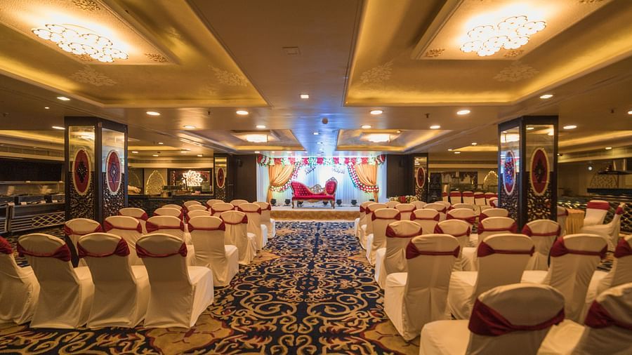 Maurya showcasing a arrangement of white-covered chairs with red sashes, a patterned carpet, and a stage area with comparable decor and lighting.
