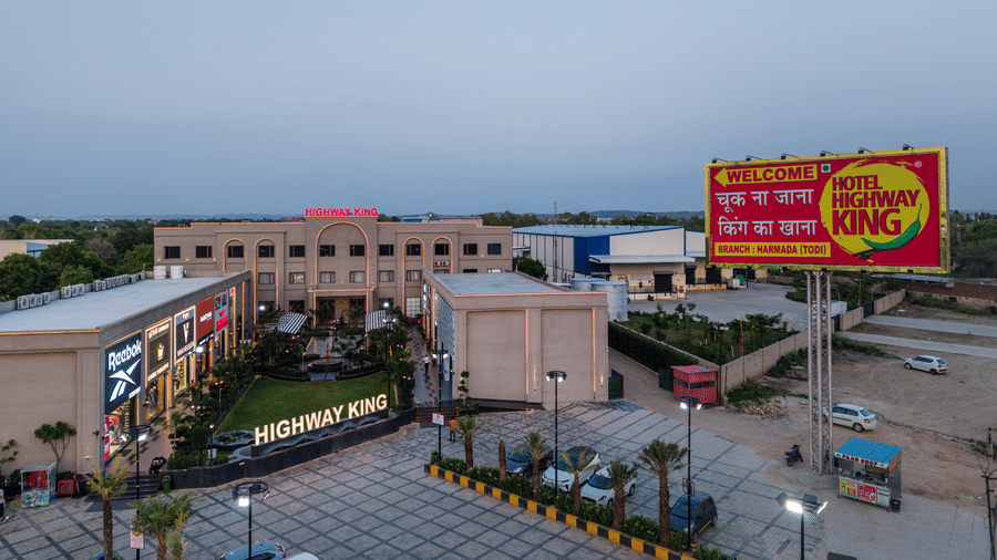 An aerial view of a large commercial complex building, a paved outdoor parking area, and a tall promotional billboard at Hotel Highway King, Harmada.