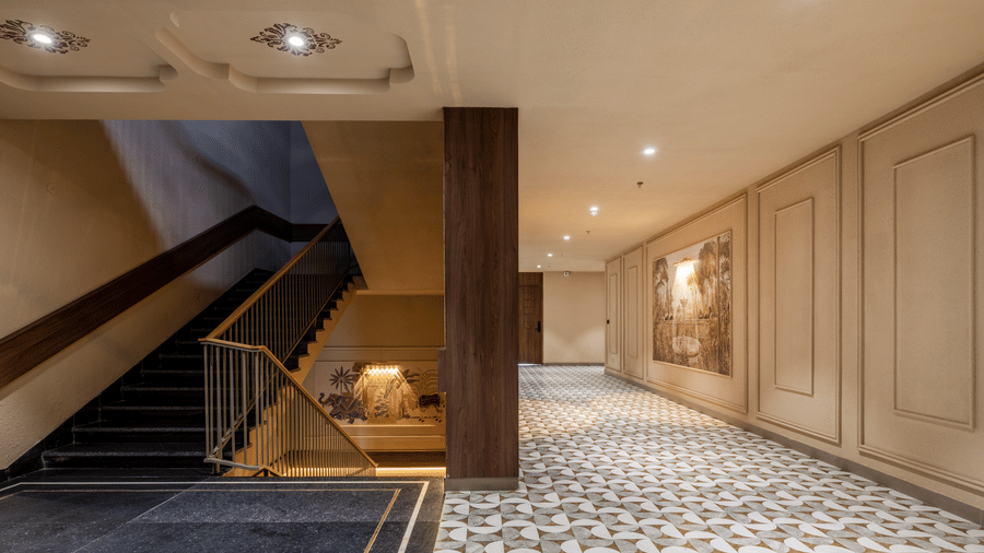 An indoor corridor area with a wide descending staircase, a dark wooden pillar support, and patterned floor tiling on the ground at Hotel Highway King, Harmada.