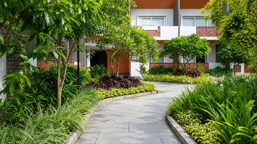 A paved walkway at Hotel Sonar Bangla, Mayapur, featuring garden plants, leafy trees, and guest room balconies.