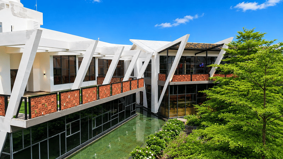 An architectural view of a building at Hotel Sonar Bangla, Mayapur, featuring slanted support beams, a balcony walkway, and a leafy tree.