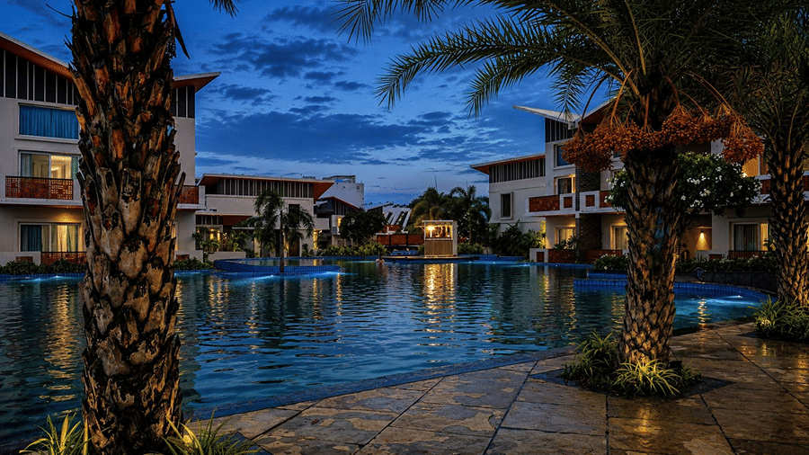 A night view of a swimming pool at Hotel Sonar Bangla, Mayapur, featuring palm tree trunks, poolside stone flooring, and guest room buildings.