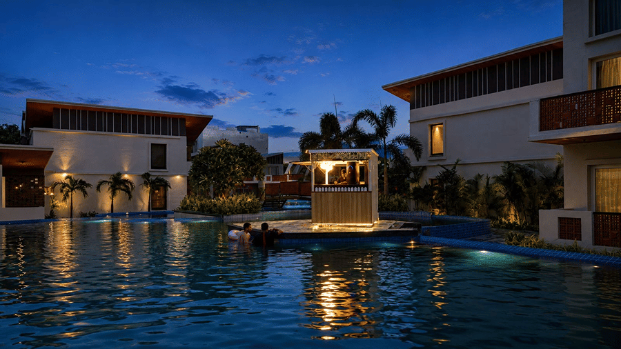 A night view of a illuminated swimming pool at Hotel Sonar Bangla, Mayapur, featuring a central wooden kiosk, palm trees, and guest blocks.