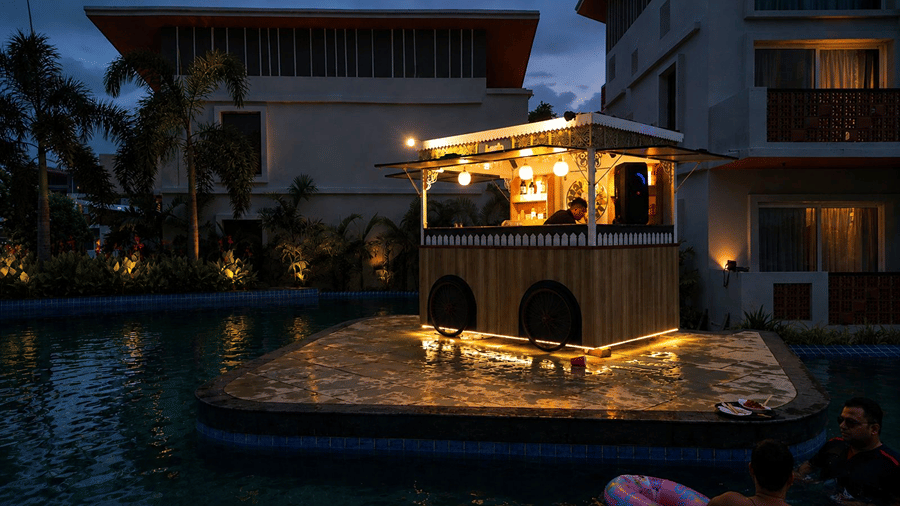 A poolside wooden kiosk structure at Hotel Sonar Bangla, Mayapur, featuring warm hanging lights, pool water, and guest room buildings.