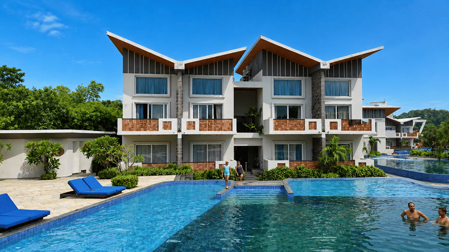 A poolside area at Hotel Sonar Bangla, Mayapur, featuring blue sun loungers, swimming pool water, guest rooms, and a clear blue sky.
