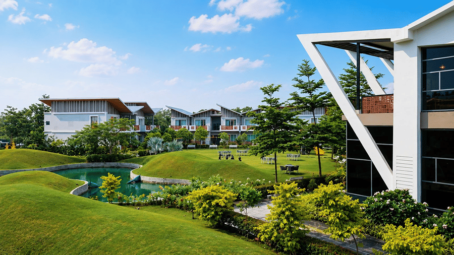 An outdoor view of Hotel Sonar Bangla, Mayapur, featuring modern building architecture, green lawns, plants, and a water pond.