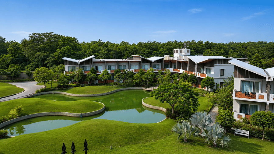 An exterior view of Hotel Sonar Bangla, Mayapur, featuring a water pond, green lawns, trees, and building structures.