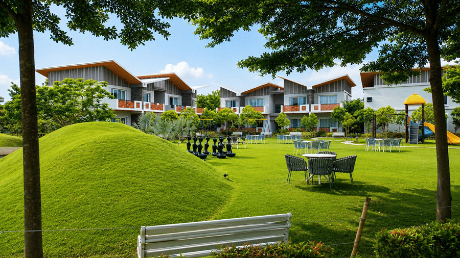 A resort garden at Hotel Sonar Bangla, Mayapur, featuring a white bench, outdoor tables with chairs, and accommodation blocks.