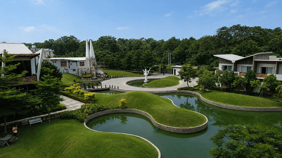 An outdoor landscape view at Hotel Sonar Bangla, Mayapur, featuring a curved water pond, grassy mounds, and resort buildings.