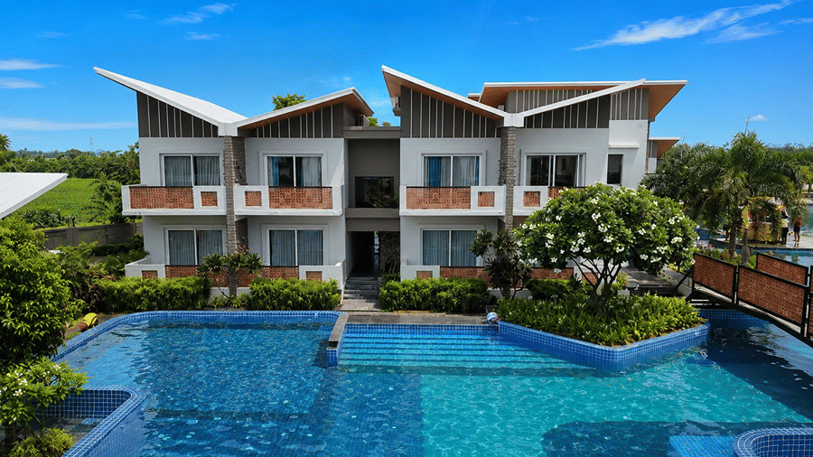 A daytime view of a swimming pool at Hotel Sonar Bangla, Mayapur, featuring a 2 storey building, glass windows, balconies, and green trees.