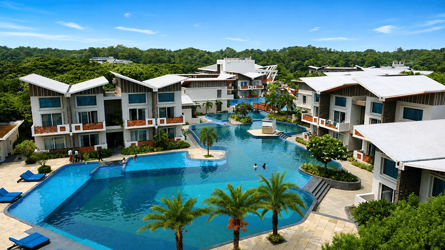 A swimming pool complex at Hotel Sonar Bangla, Mayapur, featuring outdoor sun loungers, palm trees, and multi-storey resort buildings.