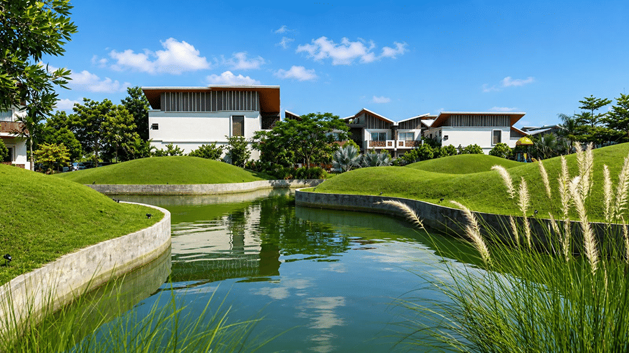 A water body at Hotel Sonar Bangla, Mayapur, featuring surrounding grass mounds, tall reed plants, and resort structures.