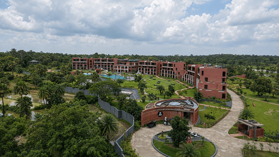 A wide aerial view of Hotel Sonar Bangla, Susunia featuring resort buildings, swimming pool, surrounding greenery, curved driveway, and lush landscape.