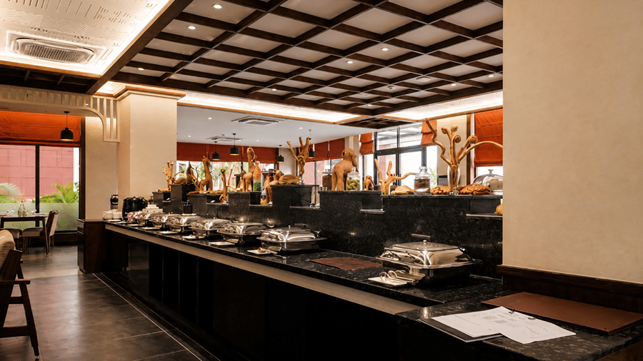 A buffet service area at Hotel Sonar Bangla, Susunia featuring stainless steel chafing dishes, food display counters, and wooden ceiling frames.