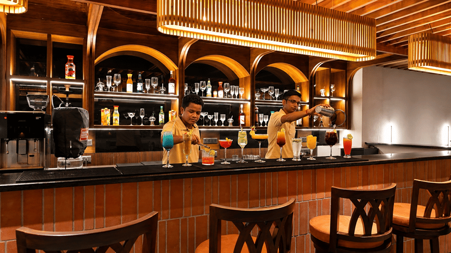 A bar counter at Hotel Sonar Bangla, Susunia featuring bartenders, mixed drink glasses, wooden bar stools, and shelf displays.   