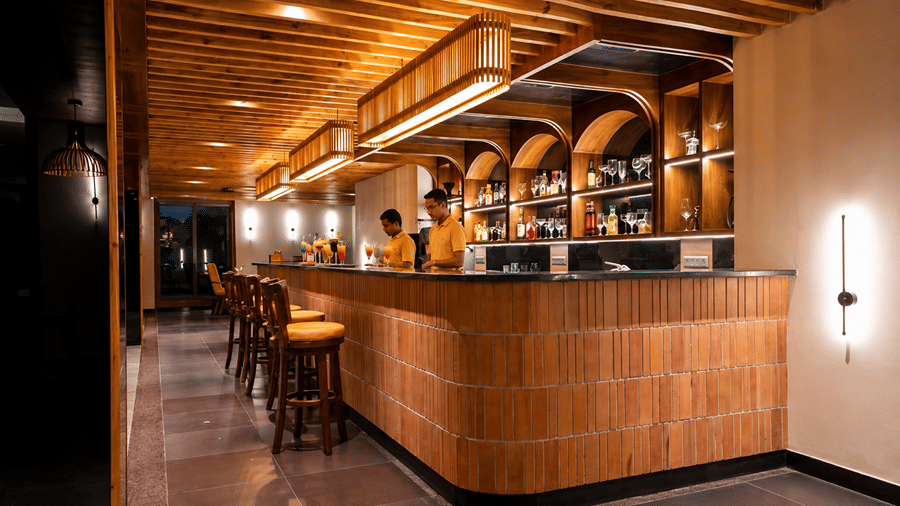 A long beverage counter at Hotel Sonar Bangla, Susunia featuring wooden bar stools, overhead lighting fixtures, and bar staff.