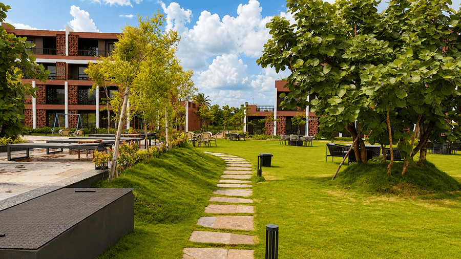A garden path at Hotel Sonar Bangla, Susunia featuring stone stepping tiles, lawn grass, outdoor seating tables, trees, and resort buildings.