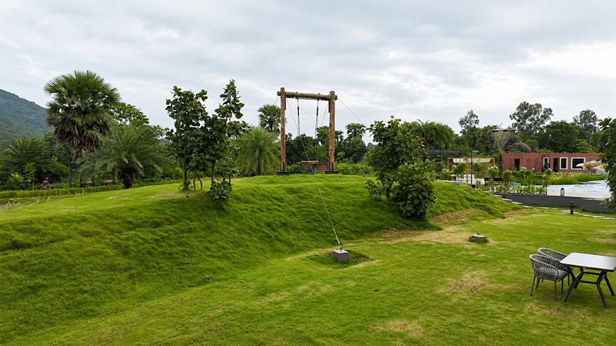 A green lawn at Hotel Sonar Bangla, Susunia featuring a tall wooden swing structure, surrounding trees, hills in the background, and outdoor seating.