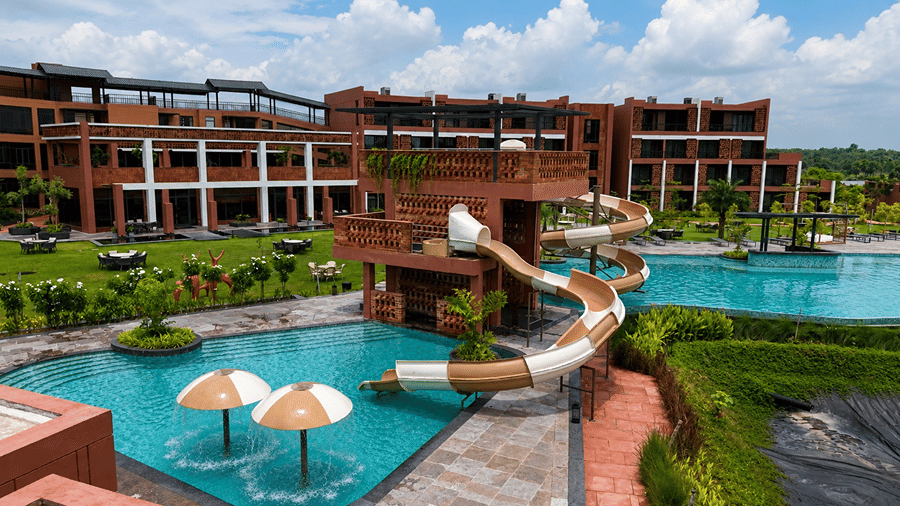 A resort pool area at Hotel Sonar Bangla, Susunia featuring a water slide tower, shallow pool with water umbrellas, and resort buildings.