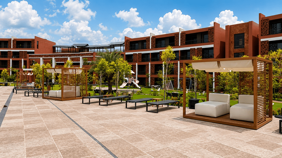 A paved outdoor terrace at Hotel Sonar Bangla, Susunia featuring wooden cabana seating, sun loungers, resort guest rooms, and garden foliage.
