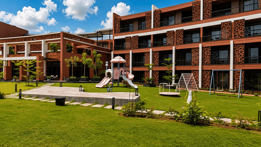 A children play area at Hotel Sonar Bangla, Susunia featuring play structures, swings, brick accommodation wings, and green lawn.