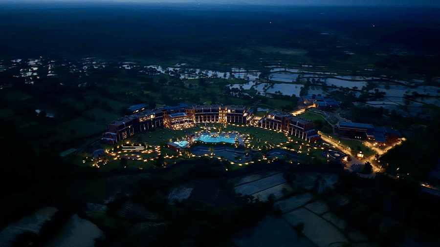 A high-angle dusk view of Hotel Sonar Bangla, Susunia featuring illuminated resort wings, lit pool, central lawns, and surrounding fields.