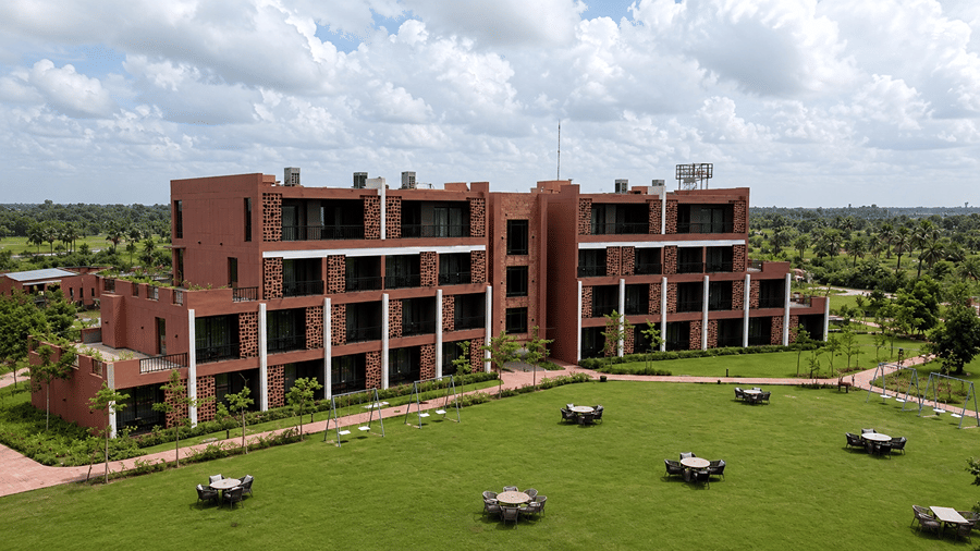 An aerial view of Hotel Sonar Bangla, Susunia featuring a multi-storey resort building, a wide green lawn, outdoor dining tables, and paved pathways.