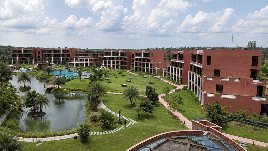 A wide garden view of Hotel Sonar Bangla, Susunia featuring a natural pond, palm trees, stone walkways, and resort accommodation wings. 