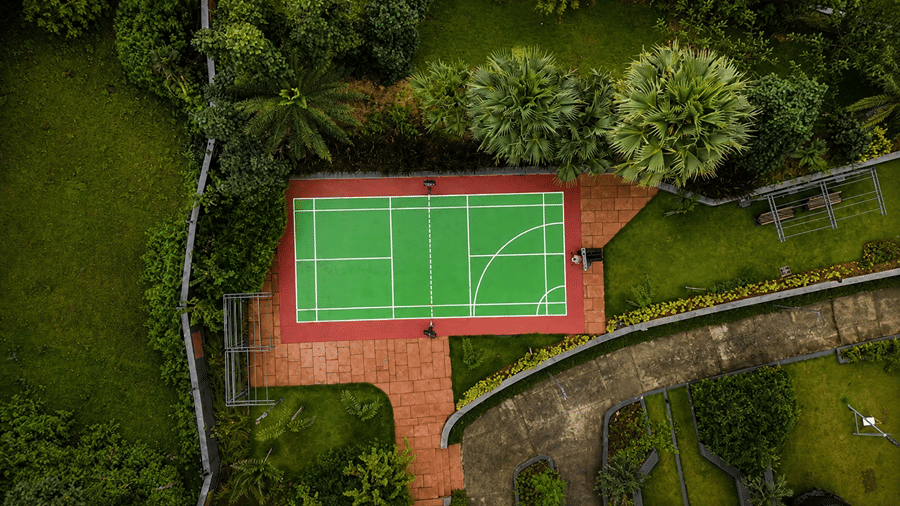 An aerial view of an outdoor sports court at Hotel Sonar Bangla, Susunia featuring marked play lines, surrounding trees, stone path, and brick paving.