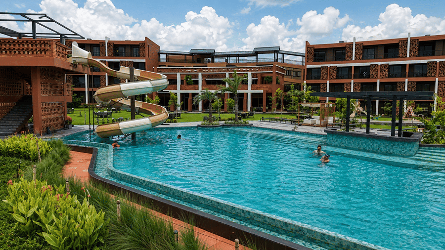 A swimming pool at Hotel Sonar Bangla, Susunia featuring a spiral water slide, surrounding resort buildings, and palm trees.