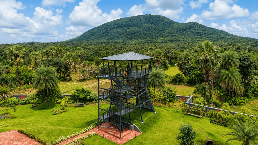 A steel watchtower structure at Hotel Sonar Bangla, Susunia featuring surrounding green lawns, forest trees, and Susunia Hill in the distance. 