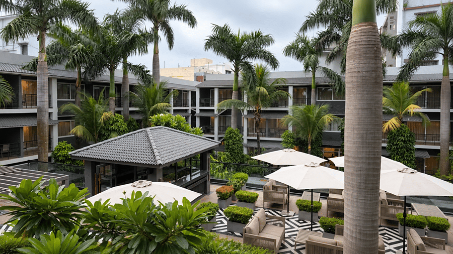 An outdoor courtyard at Hotel Sonar Bangla, Tarapith featuring patio seating, parasols, palm trees, central pavilion, and surrounding guest rooms.