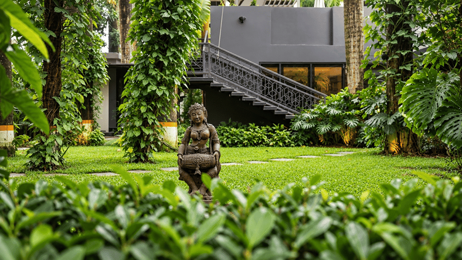 A garden area at Hotel Sonar Bangla, Tarapith featuring a stone statue, green lawn, dense trees, foliage, and outdoor staircase.