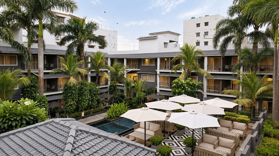 An elevated view of the cafe at Hotel Sonar Bangla, Tarapith featuring patio furniture, parasols, palm trees, pool area, and multi storey buildings.