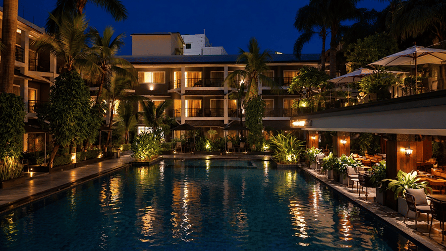 An illuminated swimming pool at night at Hotel Sonar Bangla, Tarapith featuring poolside dining tables, ambient lighting, palm trees, and guest balconies.