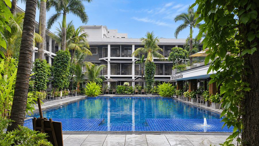 An outdoor swimming pool at Hotel Sonar Bangla, Tarapith featuring clear water, surrounding palm trees, garden plants, and hotel balcony wings.