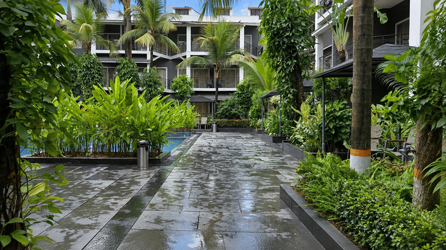A paved walkway at Hotel Sonar Bangla, Tarapith featuring adjacent swimming pool, tropical plants, palm trees, and hotel building structure.