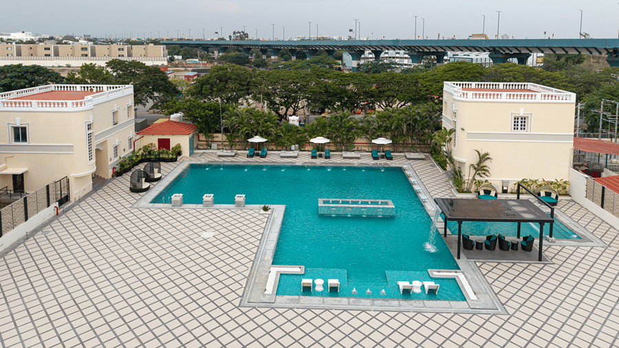 High-angle view of an L-shaped outdoor swimming pool surrounded by a spacious tiled deck and sun loungers at Jenneys Residency.
