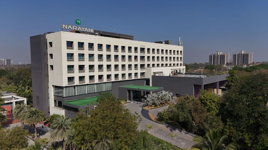 Long-shot of Narayani Heights Hotel & Resorts, Ahmedabad facade with trees surrounding it under a clear sky.