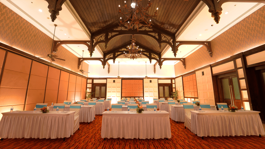 An elegant conference hall in Pondicherry at Ocean Spray, arranged in a classroom style with white skirted tables, teal chair covers, water bottles, and floral centerpieces. The venue features a carved wooden ceiling, chandeliers, and a red carpet.