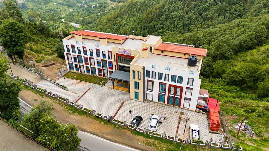 A building complex with red and white accents is seen from an elevated perspective, surrounded by lush green hills | Sukama Resort & Spa, Nainital