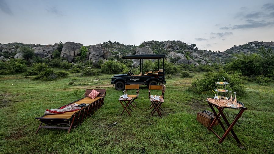An image of an outdoor seating arrangement with a jeep parked nearby amidst greenery and the backdrop of green mountains - Utsav Camp Sariska