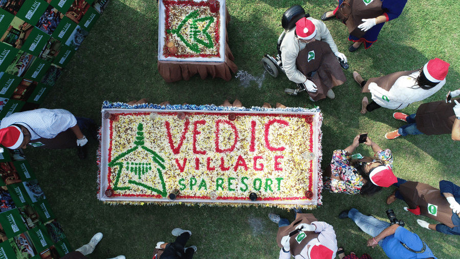 An aerial view of a floral arrangement forming the Vedic Village Spa Resort logo on a green lawn with staff members in white and red outfits standing around it.