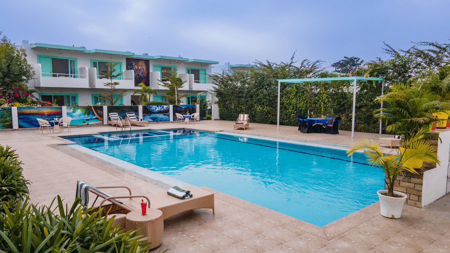 Facade of Vedikant Resorts The Mallard Corbett featuring a swimming pool, lounger beds, chairs, tables, and a two storey building with rooms.