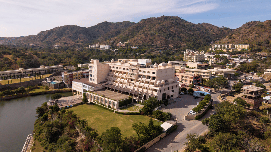 A drone view of Via Lakhela Lakeview Resort, featuring the main hotel building, landscaped lawns, a parking area, and lake beside the property.