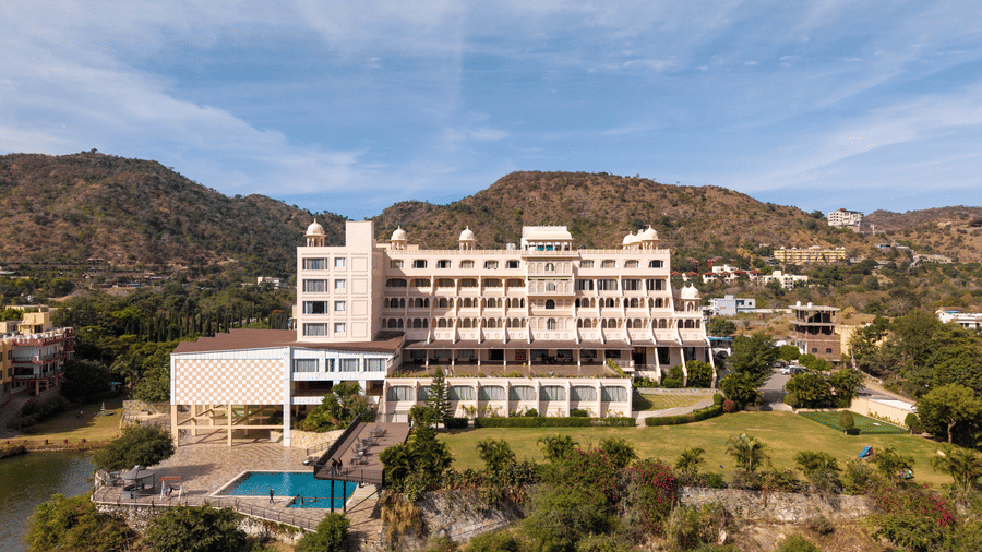 A front-facing aerial view of Via Lakhela Lakeview Resort, featuring the swimming pool, modern accommodation block, landscaped grounds, and a mountain backdrop.