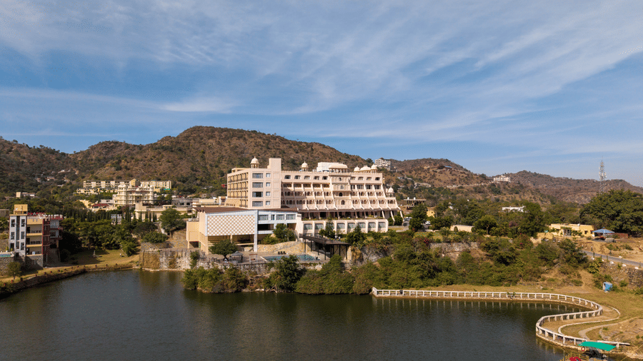 A lake-facing view of Via Lakhela Lakeview Resort, featuring the hotel facade overlooking calm waters, with rolling hills in the distance.