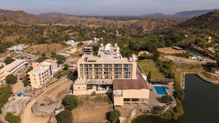 A wide drone shot of Via Lakhela Lakeview Resort, showcasing multiple resort buildings, a lakeside setting, and scenic mountain views under clear skies.